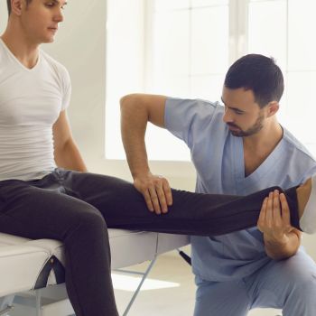 A physical therapist in blue scrubs from Middle Village Chiropractic performs a leg stretch on a seated patient in a white shirt and dark pants atop the therapy table.